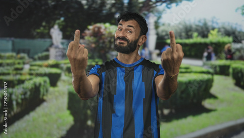 Canvastavla Bearded hispanic middle age man in blue and black striped soccer uniform shows middle fingers with bare hands in a garden setting; playful defiance