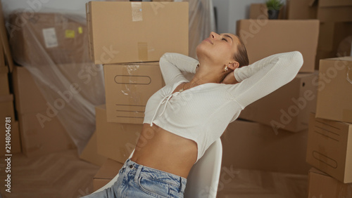 Foto Woman seated on chair stretches arms behind head showing bare midriff while surrounded by stacked moving boxes, ladder and plastic covers in a building; relief