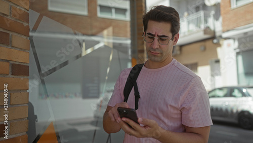 Фотография Man with glasses and backpack briefly tapping smartphone at a storefront window on a street; quiet concern