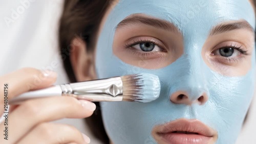 Beautiful young woman applying blue clay facial mask with a cosmetic brush. Close-up view of skincare routine, facial treatment, and spa procedure for clean skin and wellness