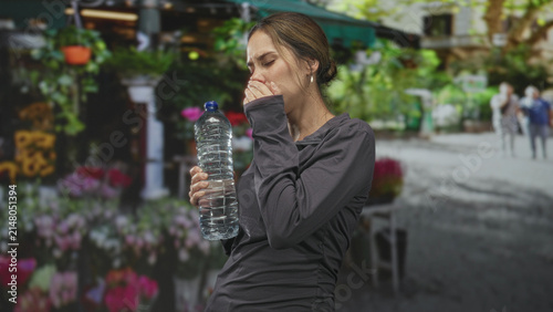 Принти на полотні Woman holding bottle covering mouth on street near flower market and potted plants; fatigue thirst relief