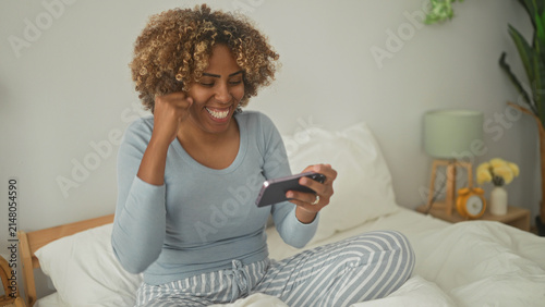 Young woman on bed in striped pajama pants holds smartphone and does a fist pump while smiling in a bright bedroom; joy celebration.