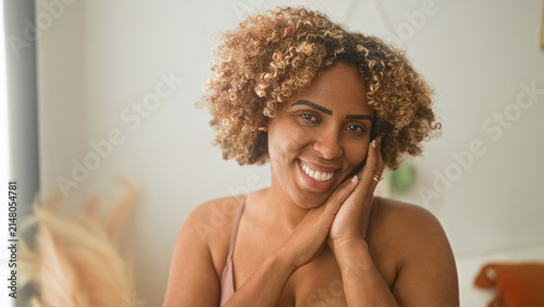 Cuadro en lienzo Woman young african american smiling with hands cupping face and clapping, bra strap and cleavage visible in studio with plant, pillow, natural curls and ring; joy