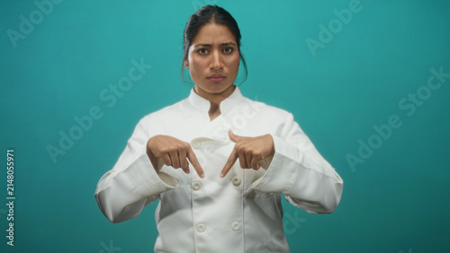 Foto Chef woman in a white jacket points both index fingers down at her chest against a teal studio backdrop; seriousness professional focus