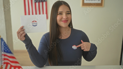 Obraz na płótnie Woman smiling holds ballot envelope and points finger to voted sticker beside american flag at polling station; civic pride