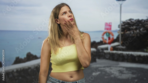 Foto Woman touching her lips with hand on a seaside street in a yellow tanktop showing midriff and jeans; pensive longing