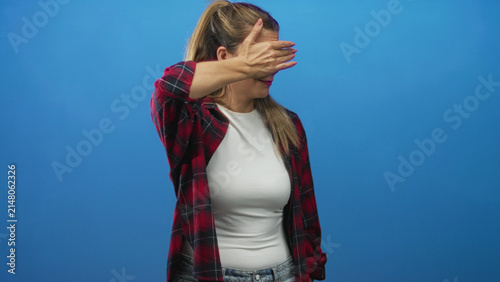 Cuadro en lienzo Woman covers eyes with hand and shields face while standing against a blue studio backdrop, wearing plaid shirt and white top; shyness privacy