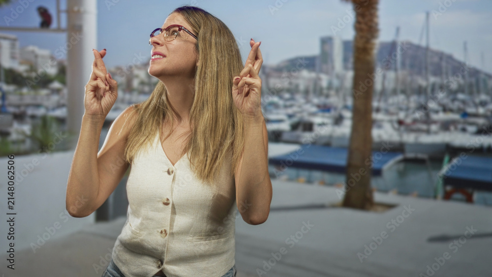 Fototapeta premium Woman with crossed fingers and glasses, hands raised on a marina street, wearing sleeveless vest; hopeful anticipation.