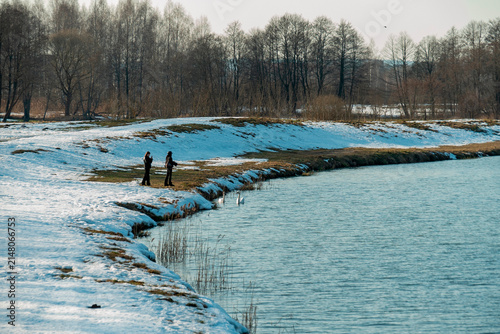 People stand by a snowy riverbank in winter as the sun shines and trees line the water's edge