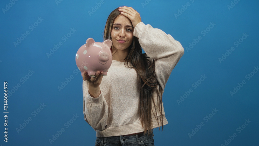 Naklejka premium Teenage woman holding pink polka dot piggy bank with hand on forehead in studio, facing camera with puzzled expression; financial worry concern.