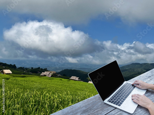 Hand typing laptop on wooden table over rice field background.