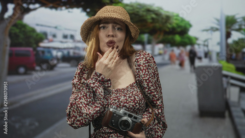 Canvas Print Woman holding vintage camera with hand to mouth gesture on a busy street wearing straw hat and floral dress; travel curiosity surprise