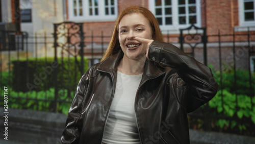 Foto Redhead woman points finger to cheek and smiles in front of building with iron fence and sidewalk; playful confidence