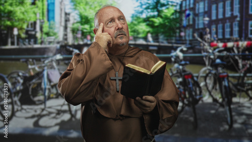 Cuadro en lienzo Man monk wearing brown robe and wooden cross holds bible and points finger to temple on street; faith reflection contemplation
