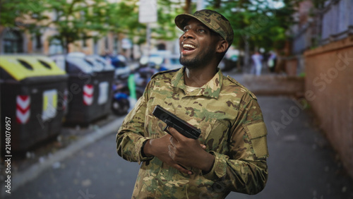 Canvastavla Soldier man in camouflage uniform holding pistol across chest, smiling and looking up amid trashbin and parked scooter on city street; pride duty