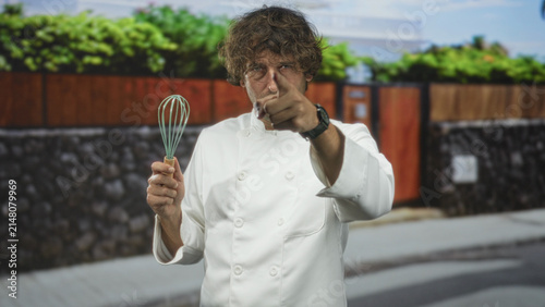 Foto Man chef in white uniform holds whisk and points index finger on street in front of stone wall and wooden fence; seriousness focus