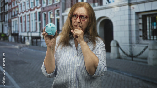 фотографія Senior woman holds blue piggy bank and presses finger to lips on a cobblestone street by a stone building; secrecy savings caution