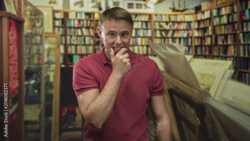 Konstfotografi Man with hand to mouth smiling and suppressing laughter while standing among crowded bookshelves in a bookstore building; amusement