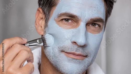 Handsome mature man applying a blue clay facial mask with a cosmetic brush. Close-up portrait of modern male skincare, spa treatment, and grooming routine for healthy, clean, and rejuvenated skin.