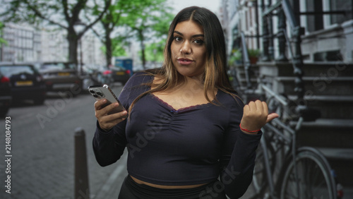 Foto Woman holding smartphone with thumb pointing to side on a city street, wearing navy top and nose ring; casual confidence