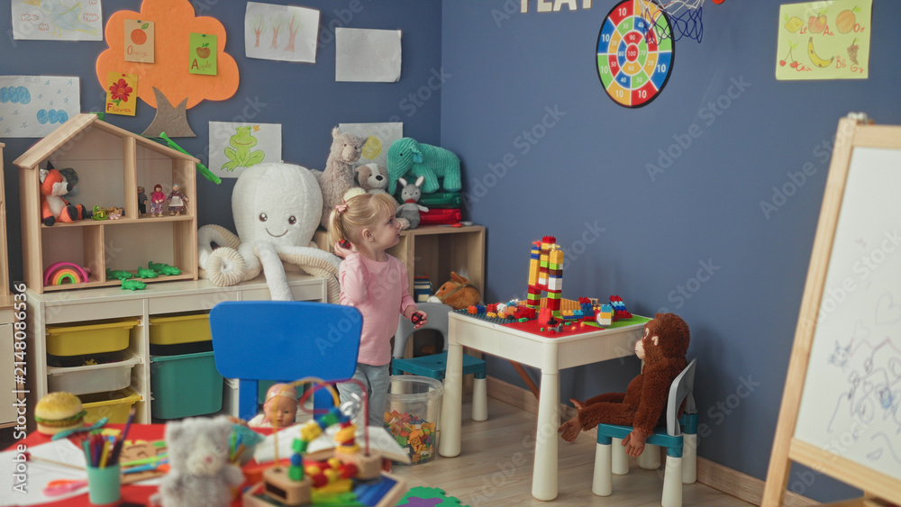 Naklejka premium Little blonde girl stacks colorful blocks at a plastic play table amid plush toys and shelving in a preschool building; curiosity wonder imagination play.