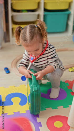 Foto Little blonde girl squats holding a green toy suitcase while wearing a red toy stethoscope and playing on foam puzzle mats in a bright playroom; curiosity