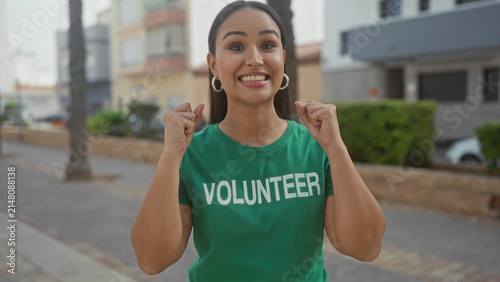 Obraz na płótnie Woman volunteer in green t shirt smiling with thumbs up and clapping while pointing at camera on street near buildings; community spirit joy