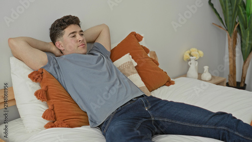 Quadro su tela Young man reclines on bed holding tv remote and points it toward the screen in a modern bedroom with pillows and plants; relaxation calm