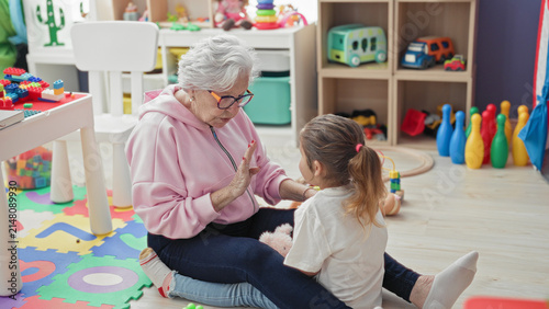 Принти на полотні Elderly woman reaches arms to hug little girl on a toy strewn playmat inside a building; affection bonding