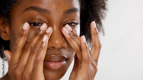 Beautiful young Black woman applying collagen under-eye patches for hydrating skincare, dark circle reduction, and anti-aging face treatment in a clean studio setting.