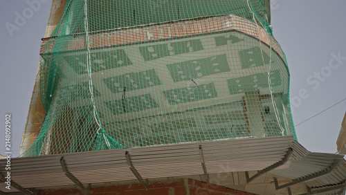 Fotografie Building balcony draped with green safety netting over unfinished brickwork, net tied to anchors and sagging along curved edge, crane top visible; caution safety