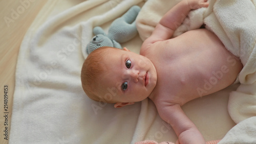 Photography Baby girl lying on soft blanket showing bare torso and raised arm in nursery with plush toys and towel; serenity innocence warmth