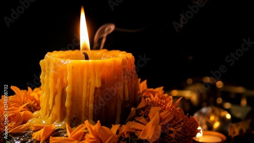 Burning beeswax candle surrounded by bright orange marigold petals with rising smoke for Day of the Dead alter decorations. Dark background with warm glowing light and copy space.
