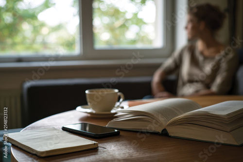 A serene indoor scene featuring a woman lost in thought while observing the view outside, surrounded by a cup of coffee, a smartphone, and an open book.
