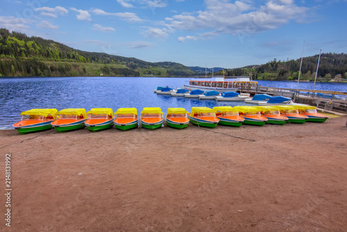 Colorful pedal boats lined up along the shore of Lake Titisee with a passenger boat, calm water, and forested hills in the Black Forest, Germany.
