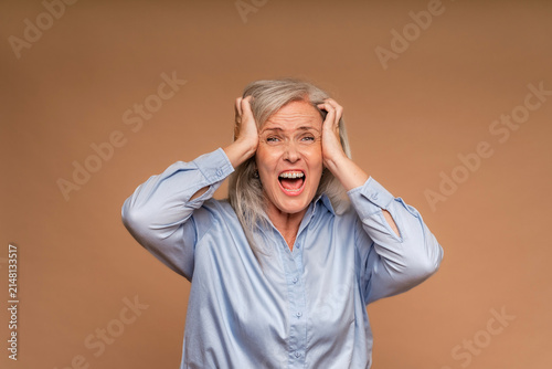 Stressed mature woman screaming in panic while holding her head on beige background