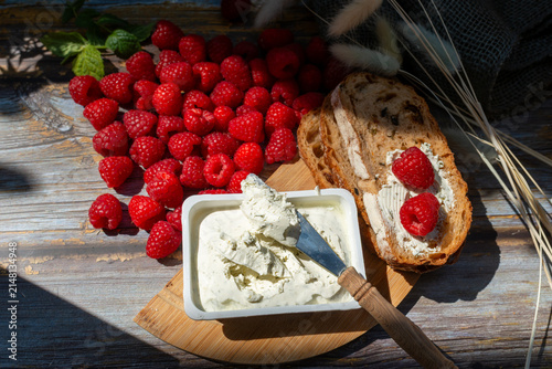 Foto Artisan bread spread with herb cream cheese and topped with ripe raspberries Rubus idaeus, served beside an open cheese container and wooden-handled knife