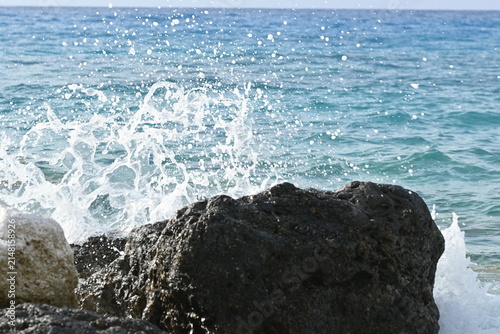 Sea water and waves splashing over a large black rock on Lefkada coast. Sparkling blue ocean under the sun with a clear horizon background, Greece.