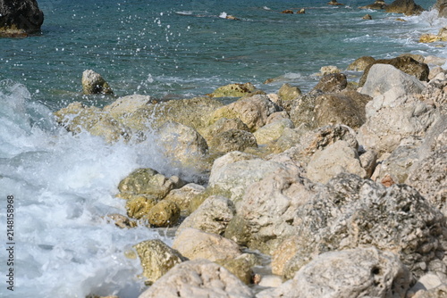 Turquoise sea and white waves washing the rocky beach of Lefkada island. Natural stone texture and crystal clear water on a sunny day in Greece.