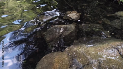 Clear lake water reflects sunlight, creating gentle ripples over submerged rocks along a natural shoreline. The peaceful movement captures the serene ambiance of a calm, pristine lake environment.
