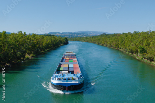 Péniche de transport de marchandises naviguant de face sur un canal entouré d'arbres.