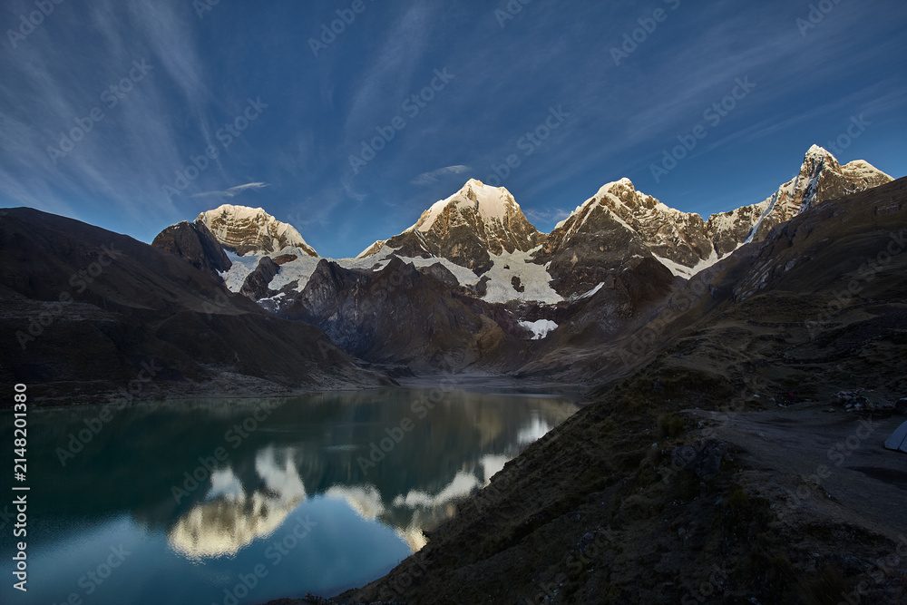 Obraz premium Beneath a dramatic sky, the snow crowned summits of Siula, Yerupajá, and Jirishanca rise in perfect formation above Carhuacocha Lagoon, their jagged silhouettes mirrored flawlessly in the still, glass