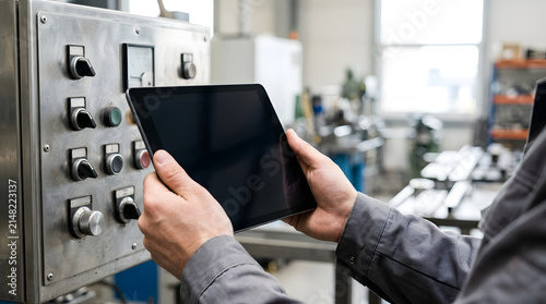 Close up of worker hands holding a blank tablet screen in front of an industrial control panel. Technician using digital device for machinery inspection in a factory