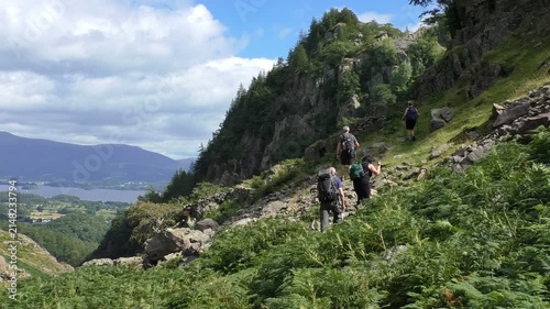 Group of four senior hikers with backpacks and walking trekking poles are climbing mountain slope in Lake District, UK Beautiful scenery with Derwent Water in the background
