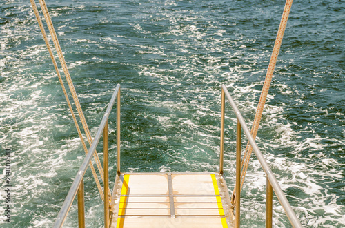 Empty cruise boat ladder and metal handrails over splashing turquoise sea water foam on a sunny summer day close up.