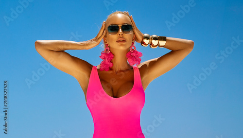 Stunning blonde woman in neon pink swimsuit and sunglasses posing at luxury tropical resort with ocean view and palms.