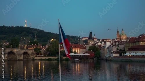 Czech national flag flutters in foreground against Prague skyline at dusk. Illuminated Charles Bridge and Vltava River reflect evening lights. Iconic Czechia architecture represents travel.