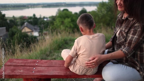 Close up slow motion shows mother gently comforting son on hilltop bench. Beautiful lake village forest and fields surround loving family moment with parental support.