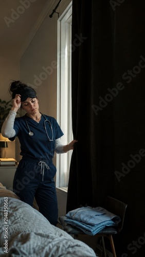 Exhausted nurse standing beside a bedroom window after a night shift, healthcare burnout awareness for social advertising