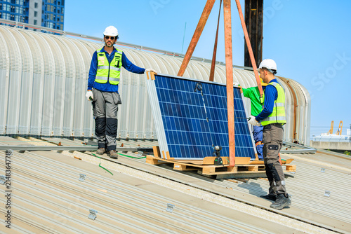 Three solar technicians in helmets and reflective vests guide a crane-lifted panel onto a metal rooftop, highlighting safe teamwork, renewable energy installation, and industrial clean power.